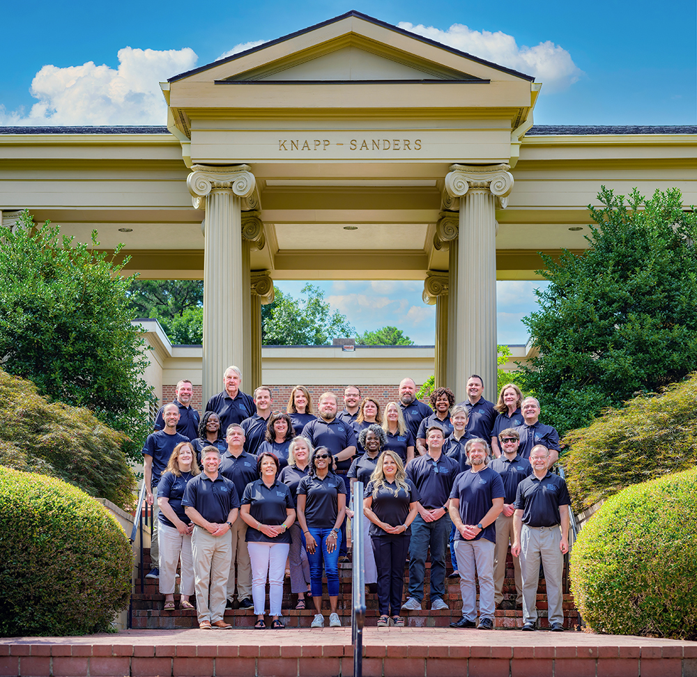 A large group of PELA graduates stand in front of a large white building with columns. They wear blue PELA polos.